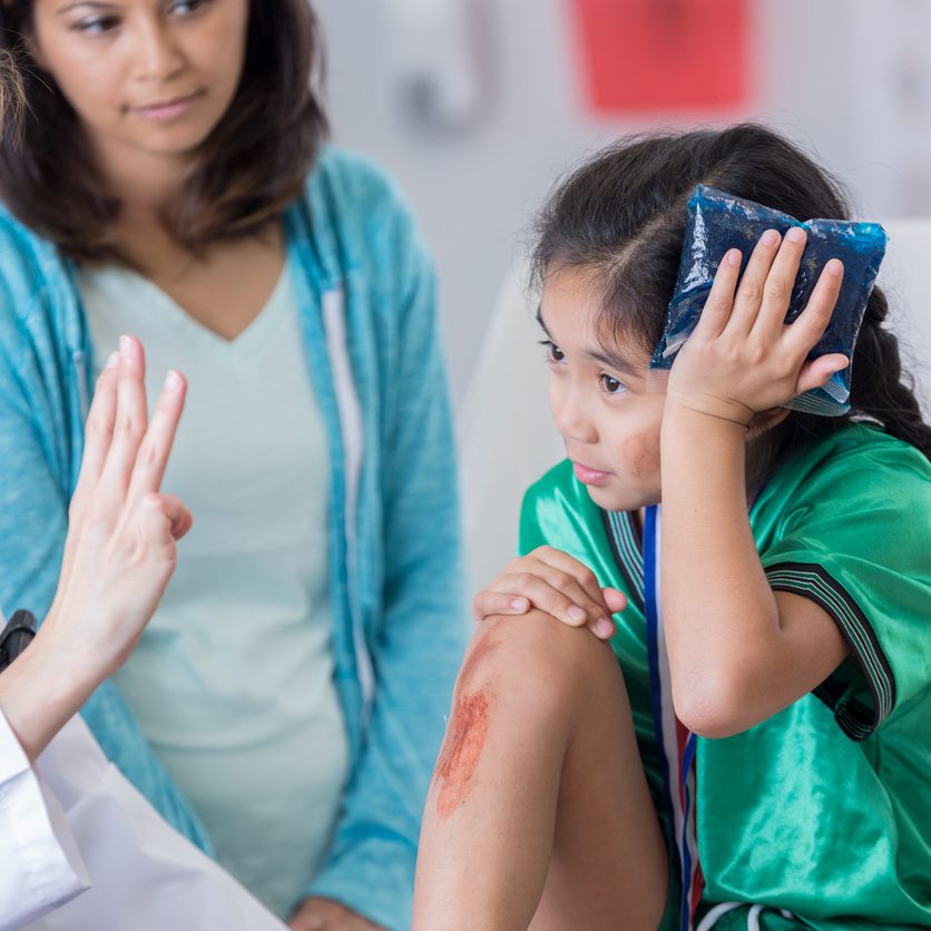 Young female soccer athlete is dazed while ER doctor asks her questions. The girl has an icepack on her head. The girl's mom is in the background.